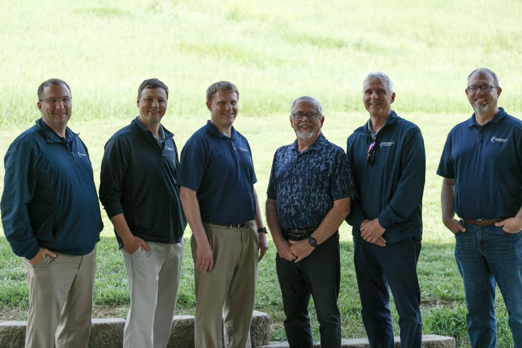 Group of six professionals in matching KC Engineering polo shirts, standing outdoors against a green backdrop, representing teamwork and collaboration in engineering and architecture careers.