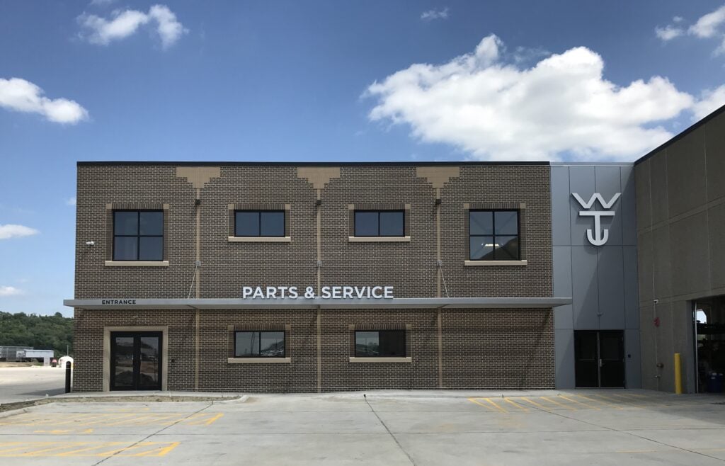 Modern building facade with "PARTS & SERVICE" signage, featuring large windows, a prominent entrance, and a logo on the side, set against a blue sky and clouds, relevant to KC Engineering's architectural design services for commercial facilities.