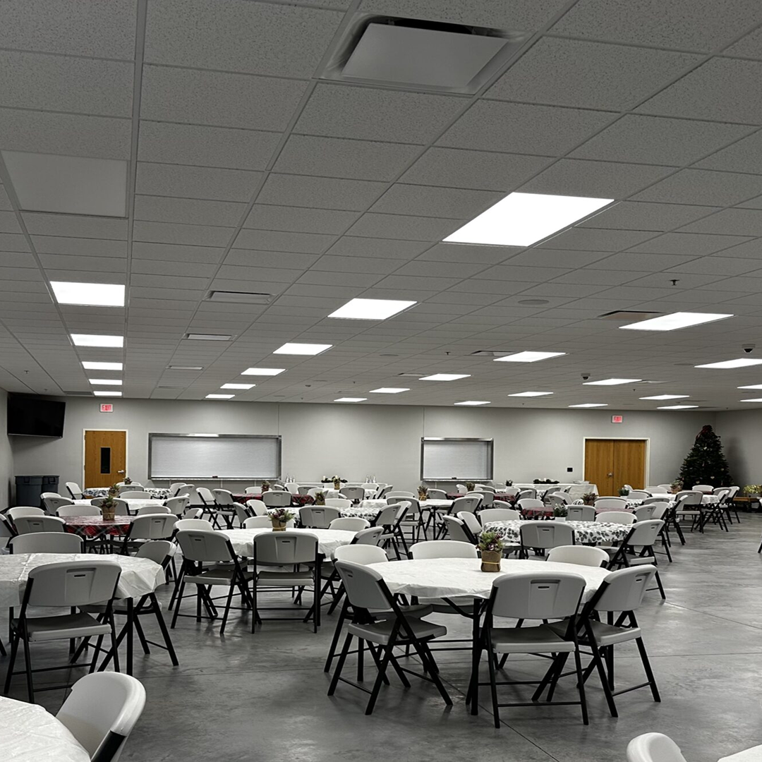 Large event space with round tables set for gathering, featuring white tablecloths and floral centerpieces, illuminated by bright overhead lights, suitable for community events or church functions.