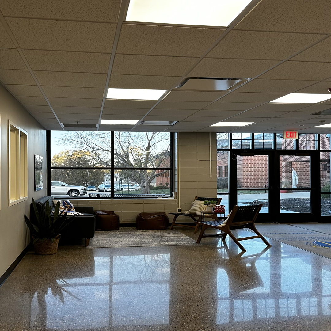 Interior view of a modern lobby with natural light, featuring a seating area with a couch, poufs, and a wooden chair, large windows providing outdoor visibility, and a welcoming atmosphere for community access and engagement.