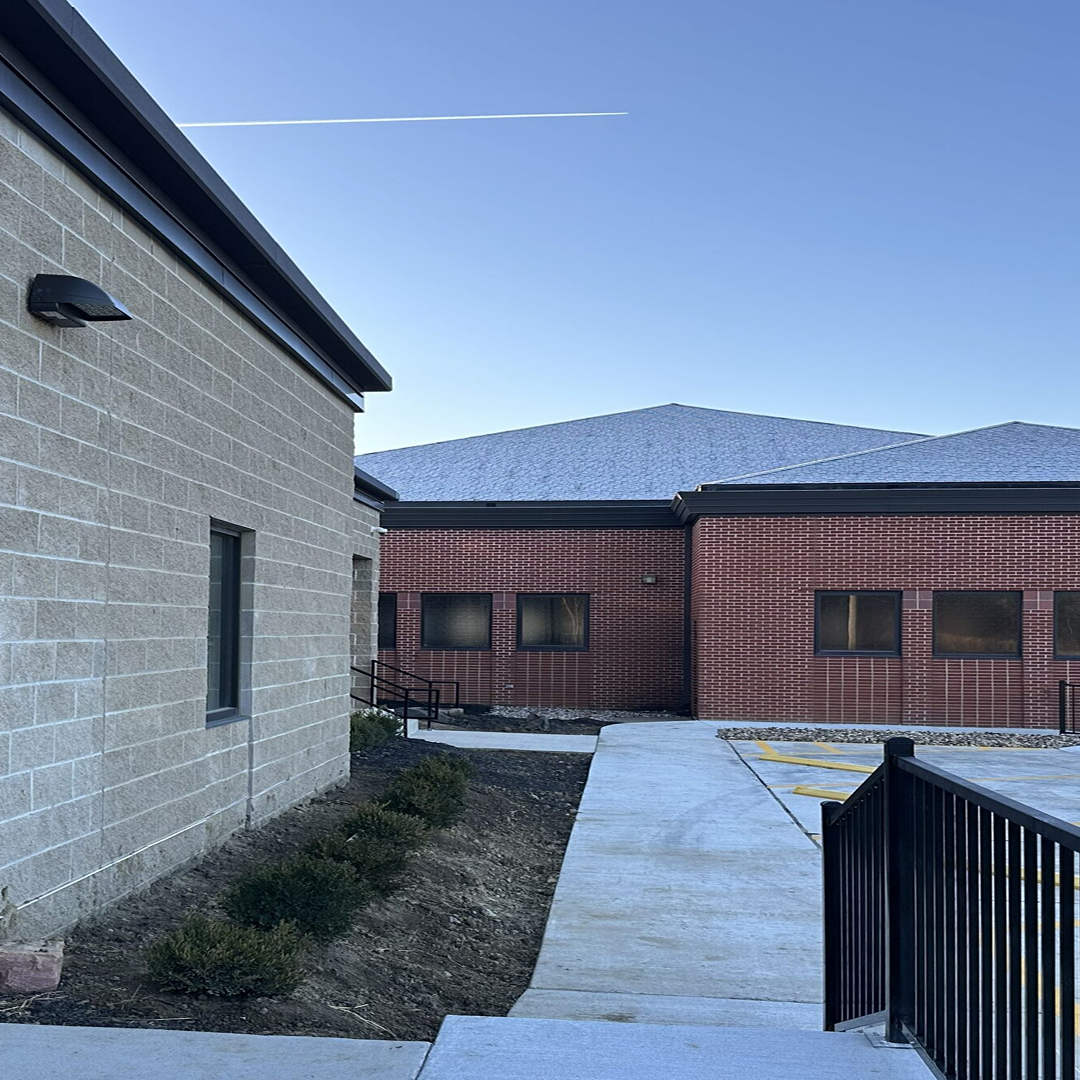 Exterior view of a modern building with a combination of textured gray block and red brick walls, showcasing a sloped roof, clear blue sky, and a pathway leading to an entrance, highlighting architectural design and accessibility features.
