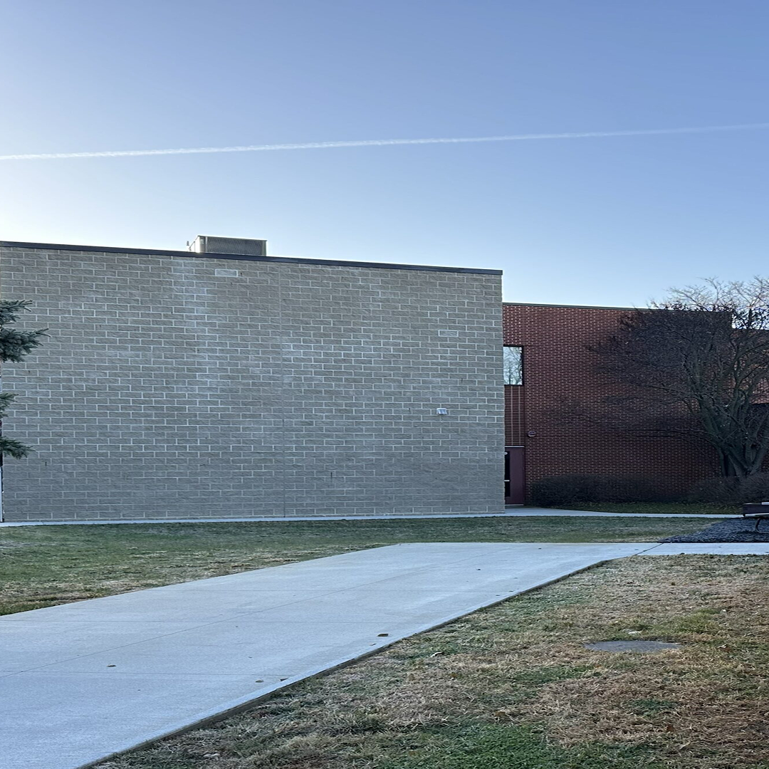 Exterior view of a building featuring a textured gray block wall and a red brick structure, with a clear sky and a pathway leading through green grass, reflecting architectural design and project planning.