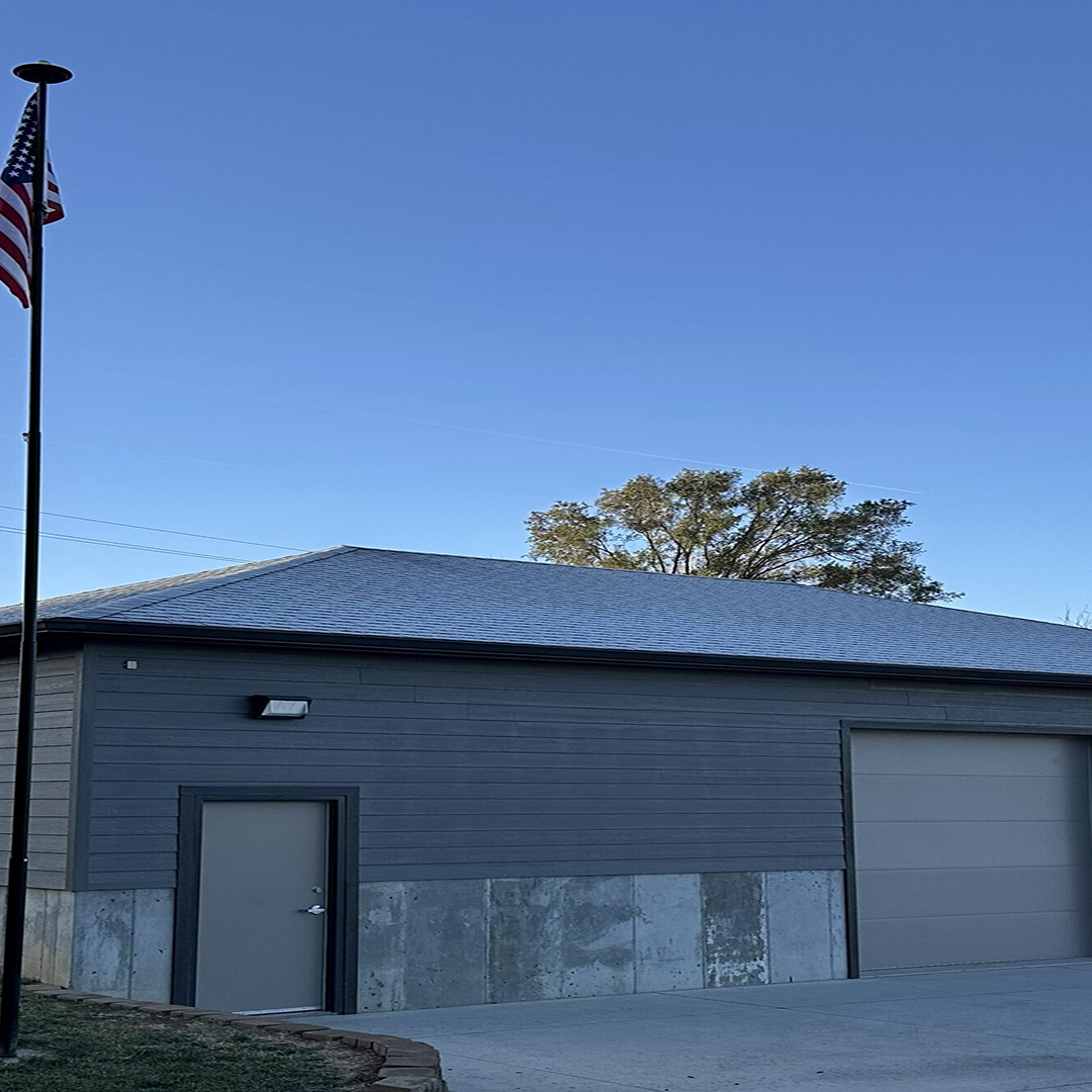 Gray building with a concrete base, featuring a door and garage door, alongside an American flag, under a clear blue sky, representing practical architectural design.