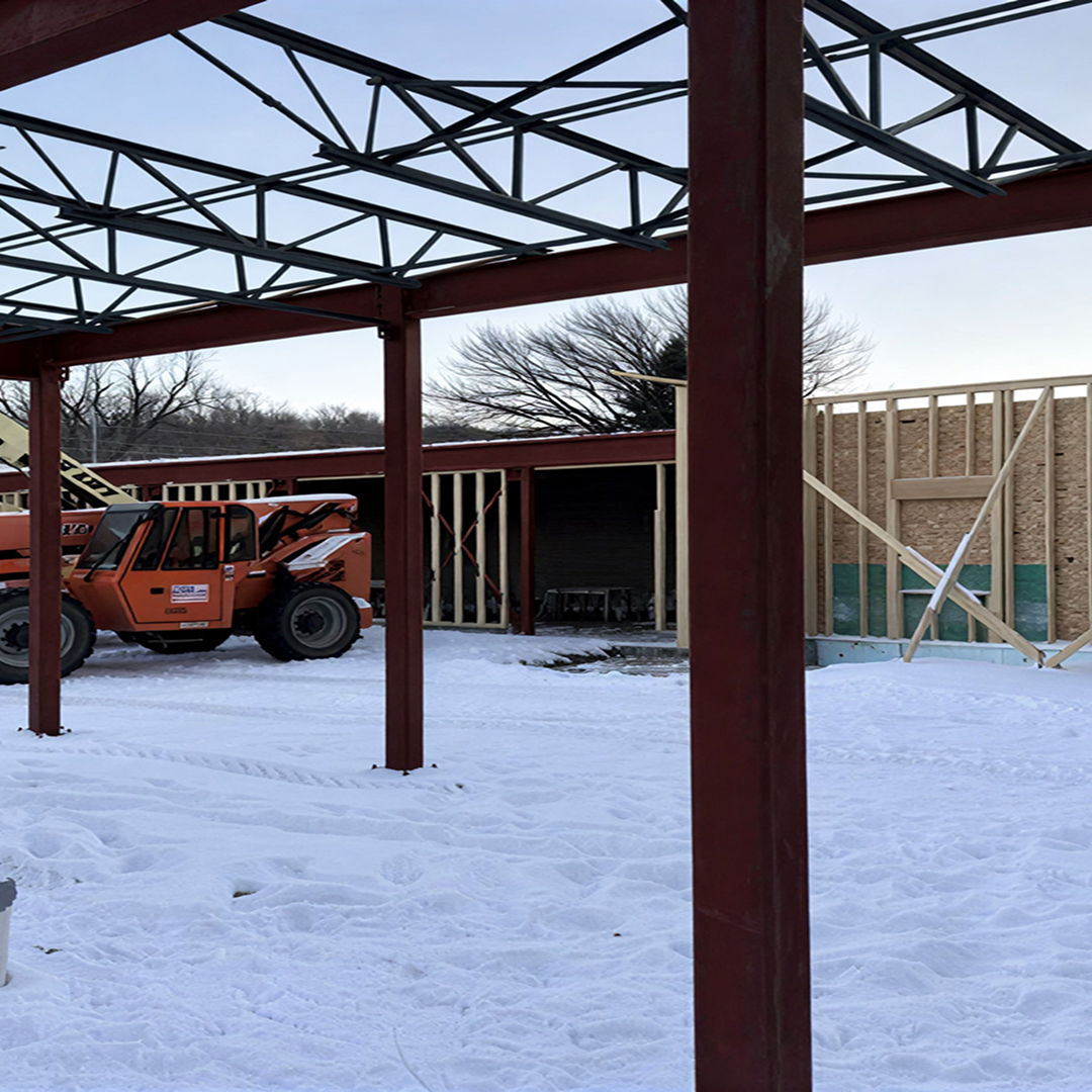 Construction site with a telehandler and framing structure, showcasing early project development in a snowy environment, emphasizing architectural and structural coordination.