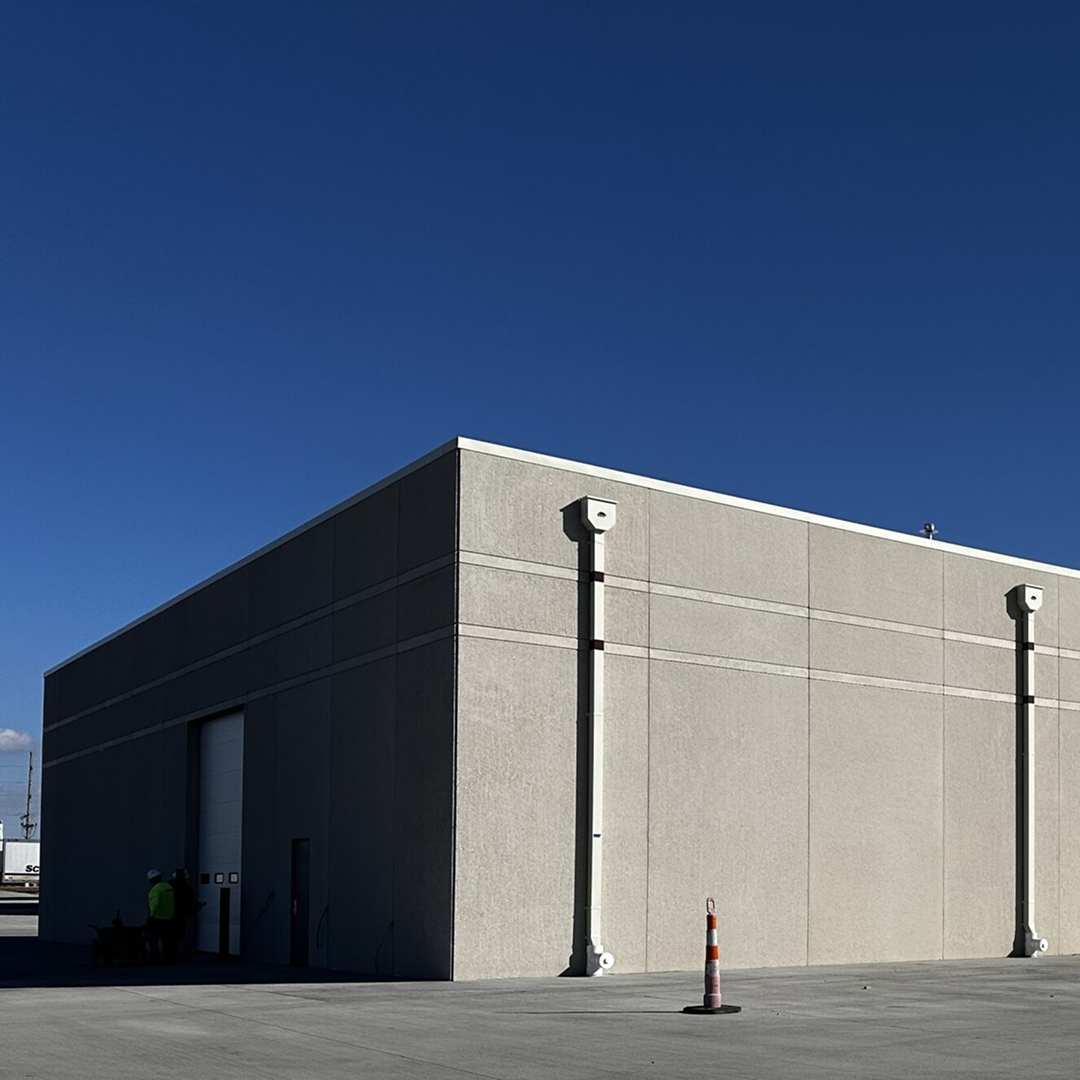 Industrial facility exterior with clear blue sky, featuring a simple structure, loading dock, and two workers in safety gear discussing project details.