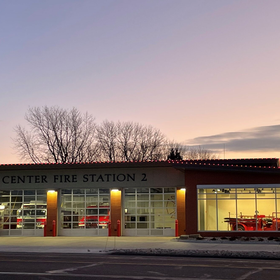 Sioux Center Fire Station 2 building exterior at dusk, featuring large glass garage doors showcasing fire trucks, surrounded by trees and a gradient sky, emphasizing community safety and architectural design.