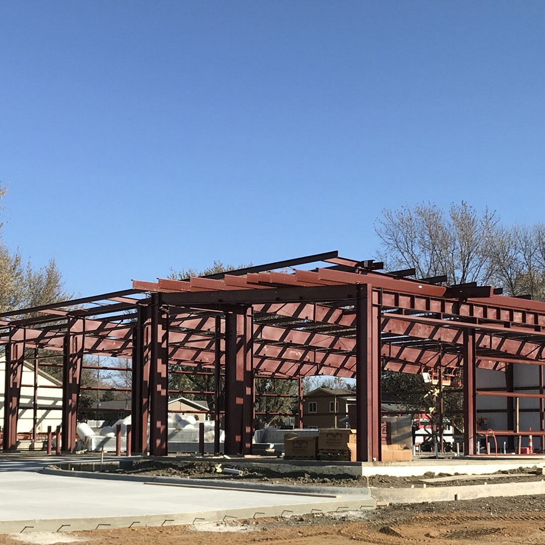 Steel framework of a fire station under construction, showcasing structural elements and foundation work, with surrounding trees and residential buildings in the background.