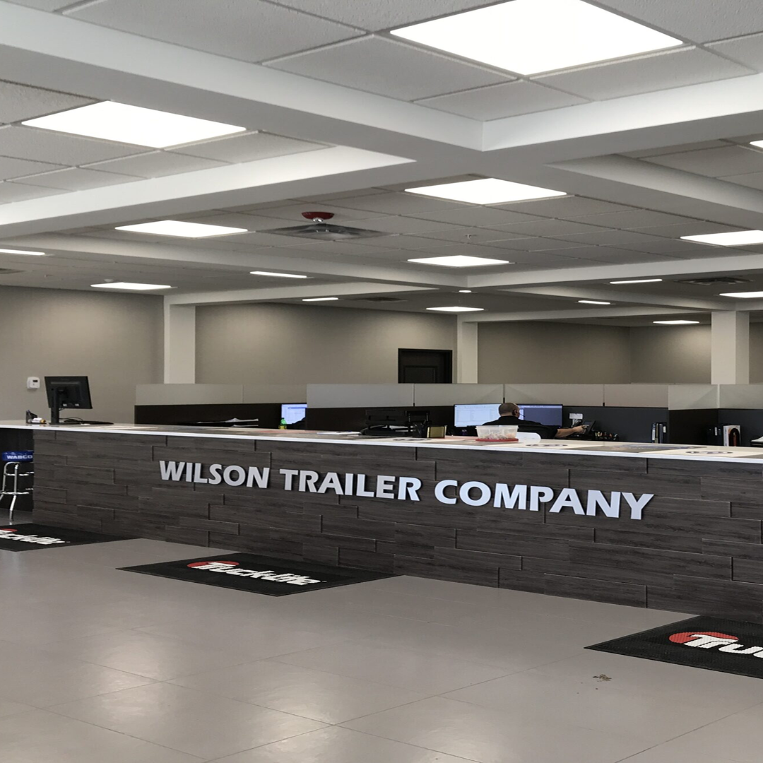 Wilson Trailer Company reception area with modern design, featuring a stone-textured counter, company name prominently displayed, and staff working at desks in the background.